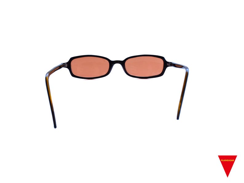May include: Oval-shaped sunglasses featuring a dark brown tortoiseshell frame and tinted orange lenses. The glasses have thin, curved temples and a bridge connecting the two rims. The design is simple with a smooth finish on the frame edges.