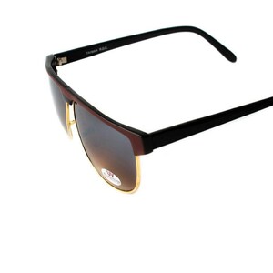 May include: Black sunglasses with brown gradient lenses and a gold trim. The sunglasses have a black frame and a black plastic arm.