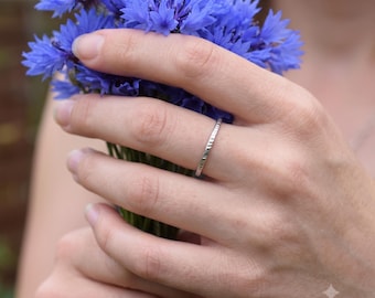 Op de afbeelding: Een dunne, zilverkleurige metalen ring met een getextureerde, gehamerde oppervlakteafwerking. De band is voorzien van een herhalend patroon van kleine, verticale inkepingen langs de gehele omtrek, wat zorgt voor een subtiele, lichtreflecterende uitstraling.