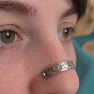 May include: A silver-toned nose bridge ring featuring a hammered, textured surface. The piece is a flat, horizontal band that rests across the bridge of the nose, secured by a small, polished spherical bead at the visible end of the metal strip.