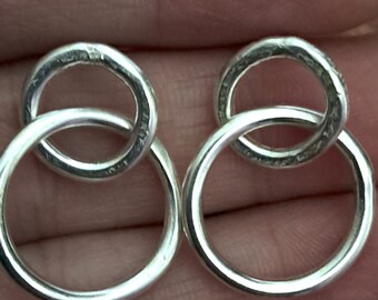 May include: A pair of silver earrings featuring a double-circle design. Each earring has two interlocking rings, one larger and one smaller. The earrings are made of a shiny, reflective metal.