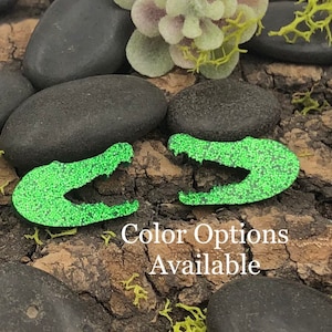 Pode incluir: Um par de brincos de cabeça de jacaré com glitter verde. Os brincos são em forma de cabeça de jacaré com a boca aberta. Os brincos são feitos de glitter e têm um acabamento brilhante.