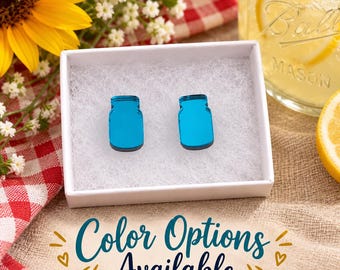 May include: A pair of bright blue, reflective, mason jar-shaped stud earrings resting inside a white gift box filled with white padding. The earrings feature a smooth, glossy finish and a distinct silhouette resembling a classic glass jar.