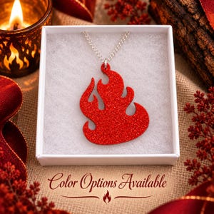 May include: A red glittery flame-shaped pendant suspended from a silver-toned chain, presented inside a white square box. The text Color Options Available is printed in a dark script font below the box, accompanied by a small flame icon.