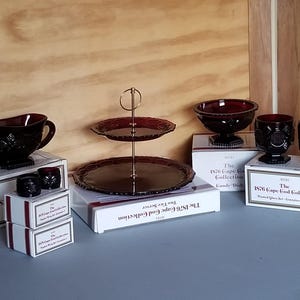 May include: A collection of ruby red glass items from the Avon 1876 Cape Cod Collection, including a gravy boat, a two-tier server with a gold-tone metal handle, a candy dish, two glasses, and two napkin rings, all displayed with their original boxes.
