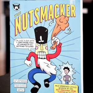 May include: A comic book cover featuring a cartoon character called "The Nutsmacker" in a red and white suit, holding a large nutcracker. The background is blue with yellow text and a white border. The text on the cover reads "The Nutsmacker" and "I'm just a guy with a pantyhose full of walnuts and nothing to lose!"  The text at the bottom reads "Mild-mannered office worker by day, nut-smacking crime fighter by night!"