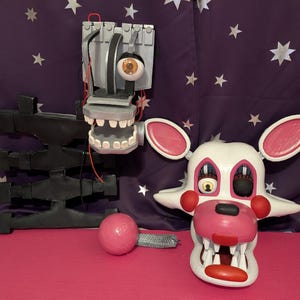 May include: A white and pink animatronic mask with large ears, red cheeks, and a wide, toothy grin. Beside it is a black and gray robotic arm and head with a single eye. A pink ball and metal piece are also present.