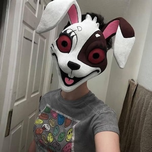 May include: A white and brown rabbit mask with large ears, red eyes, and a wide grin. The mask has black accents and is worn by a person wearing a gray t-shirt with a colorful smiley face design.