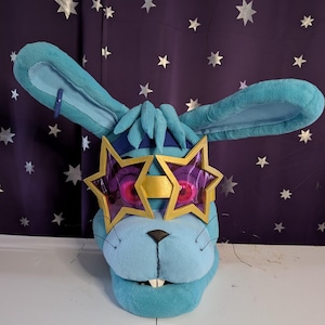 May include: A plush blue bunny head with large ears and a black nose. The bunny wears star-shaped glasses with purple lenses and a gold frame. The background is a dark purple fabric with silver star shapes.