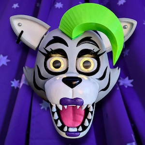 May include: A white and gray animatronic animal mask with a green mohawk. The mask has a wide open mouth with sharp teeth and a pink tongue. The eyes are yellow with black pupils.