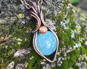 May include: A copper pendant necklace with a light blue gemstone. The gemstone is held in a wire-wrapped copper setting, with a small copper bead above it. The necklace hangs against a mossy, textured background. The pendant is a unique piece of jewellery.