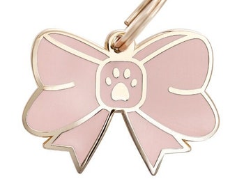 May include: A pink bow-shaped dog tag with a gold paw print in the center. The tag has a gold ring for attaching to a collar.