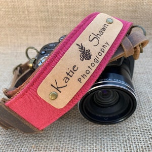 May include: A red camera strap with a brown leather trim and two personalized wooden name tags. The name tags read "Katie" and "Shawn Photography".