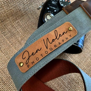 May include: A gray canvas camera strap with a brown leather patch that says "Jen Molen Photography" in black lettering.