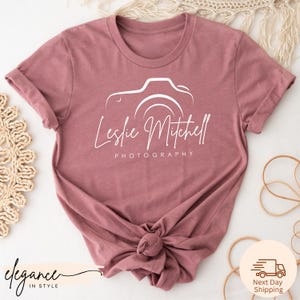 May include: Dusty rose t-shirt featuring a white camera graphic with the name "Leslie Mitchell" and the word "Photography" printed below. The shirt has rolled-up sleeves and a knotted hem. The shirt is made of soft material.