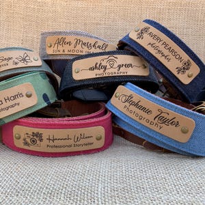 May include: A collection of six camera straps with personalized wooden nameplates. The straps are made of denim and canvas in various colors, including blue, green, red, and black. The nameplates feature different names and titles, such as "Victoria Harris, VH Photography" and "Ashley Green, Photography".