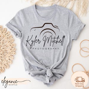 May include: A light gray t-shirt featuring a black camera design with the name "Kyler Mitchell" and the word "Photography" printed below. The shirt is knotted at the waist.