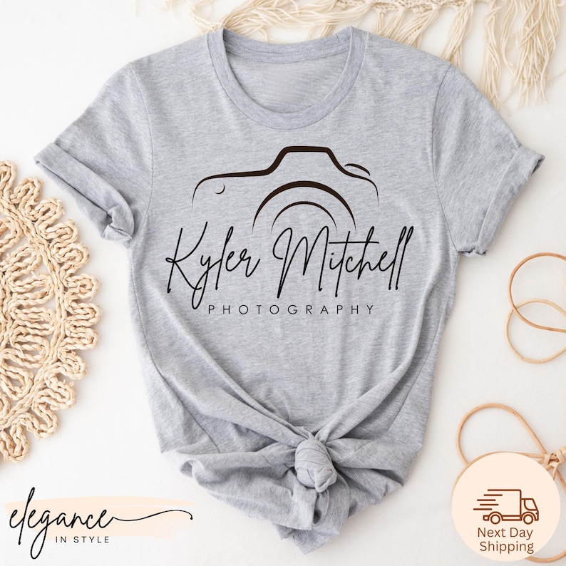 May include: A light gray t-shirt featuring a black camera design with the name "Kyler Mitchell" and the word "Photography" printed below. The shirt is knotted at the waist.