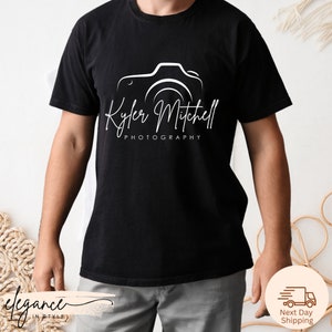 May include: Black t-shirt with a white outline of a camera and the text "Kyler Mitchell Photography" in a script font. The shirt is a crew neck style.