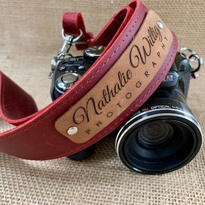 May include: A red leather camera strap with a brown leather patch that says "Nathalie Wittig Photography" in a cursive font.