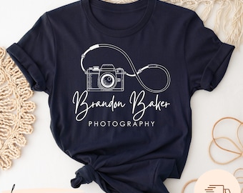 May include: A navy blue t-shirt with a white outline of a camera and a strap forming an infinity symbol. The text "Brandon Baker PHOTOGRAPHY" is written below the camera.