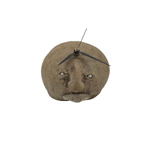 Pode incluir: Um relógio peculiar em forma de rosto com bigode. O relógio é de cor castanho claro com ponteiros pretos e olhos brancos. Um item decorativo.