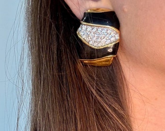 May include: A pair of black and gold earrings with a large, clear rhinestone cluster in the center. The earrings are shaped like a curved rectangle.