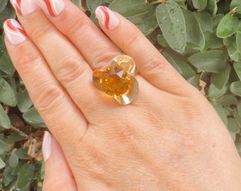 May include: A large, heart-shaped ring with a golden-yellow gemstone. The ring is set on a hand with manicured nails, against a backdrop of green foliage. The gemstone has multiple facets, reflecting light.