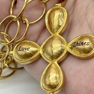 May include: Gold-toned necklace with a cross pendant. The cross is made of five round, textured pieces. The top piece has a small loop for attaching the chain. The bottom piece has a small ball. The cross has the words "Love" and "Shines" engraved on it.