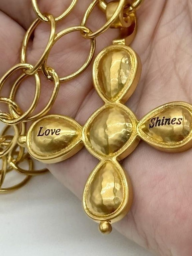 May include: Gold-toned necklace with a cross pendant. The cross is made of five round, textured pieces. The top piece has a small loop for attaching the chain. The bottom piece has a small ball. The cross has the words "Love" and "Shines" engraved on it.