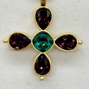 May include: A gold-tone cross pendant featuring five faceted gemstones. The central stone is a large, emerald green gemstone. The four surrounding stones are smaller, teardrop-shaped, and a deep purple colour.
