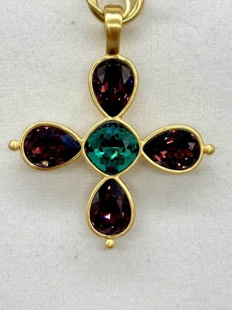 May include: A gold-tone cross pendant featuring five faceted gemstones. The central stone is a large, emerald green gemstone. The four surrounding stones are smaller, teardrop-shaped, and a deep purple colour.