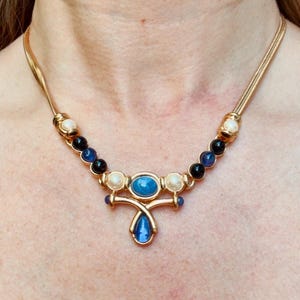 May include: Gold-tone necklace featuring a central pendant with a blue oval stone and a teardrop-shaped blue stone. The necklace has a chain design with alternating black, blue, and white beads. The necklace is approximately 16 inches long.