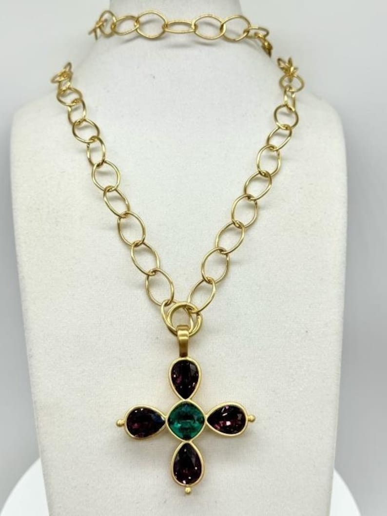May include: A gold chain necklace with a pendant in the shape of a cross. The cross is adorned with five faceted gemstones: one large green stone in the centre and four smaller purple stones around it.