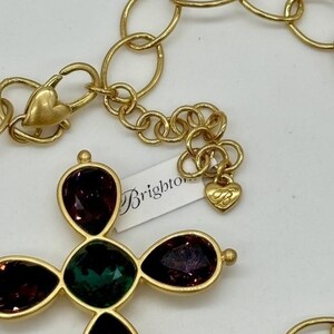 May include: Gold chain necklace with a large cross pendant featuring green and purple gemstones. The pendant is attached to a gold chain with a heart-shaped clasp. The necklace has a "Brighton" tag attached.