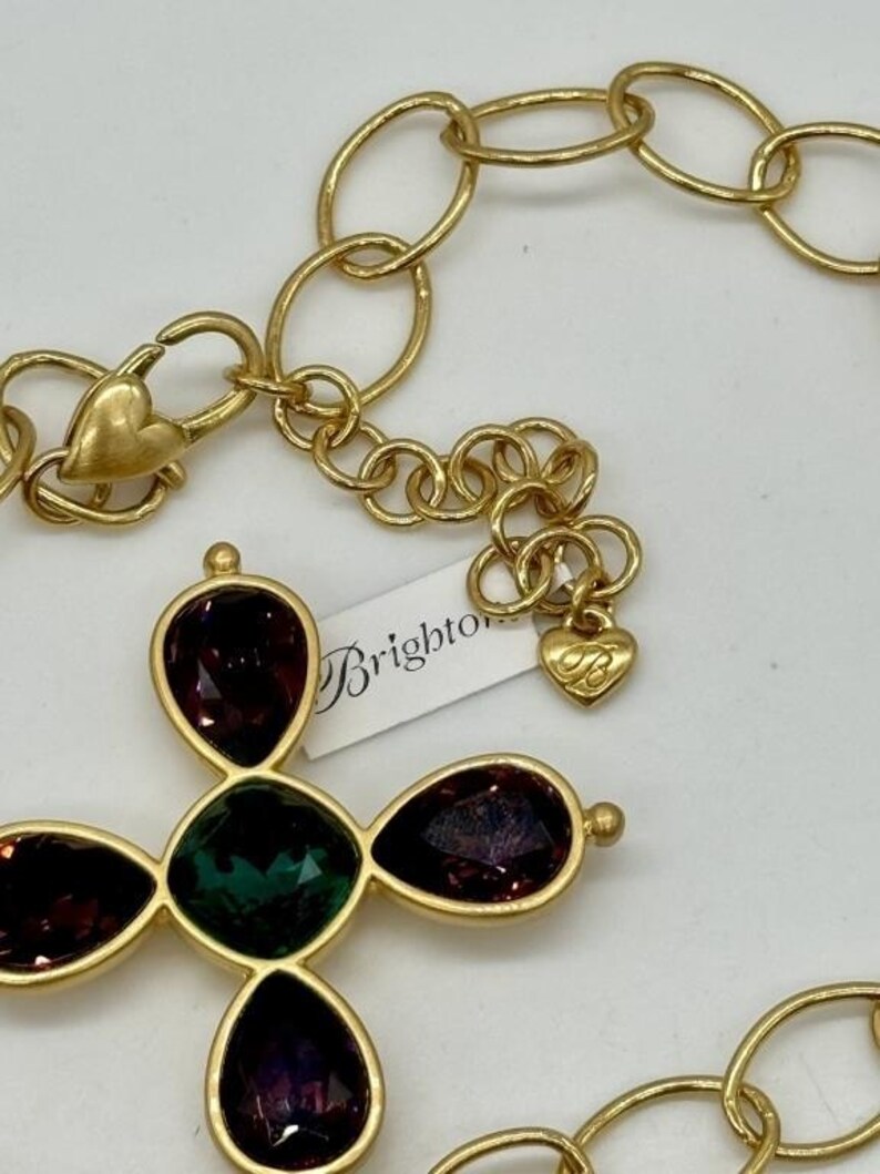 May include: Gold chain necklace with a large cross pendant featuring green and purple gemstones. The pendant is attached to a gold chain with a heart-shaped clasp. The necklace has a "Brighton" tag attached.