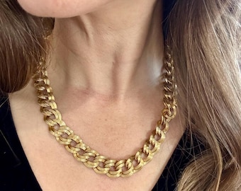 Puede incluir: Un collar de cadena dorado con un diseño texturizado y enlazado. El collar es de estilo grueso, con cada eslabón que parece estar hecho de múltiples hebras. El collar se usa sobre una blusa negra.