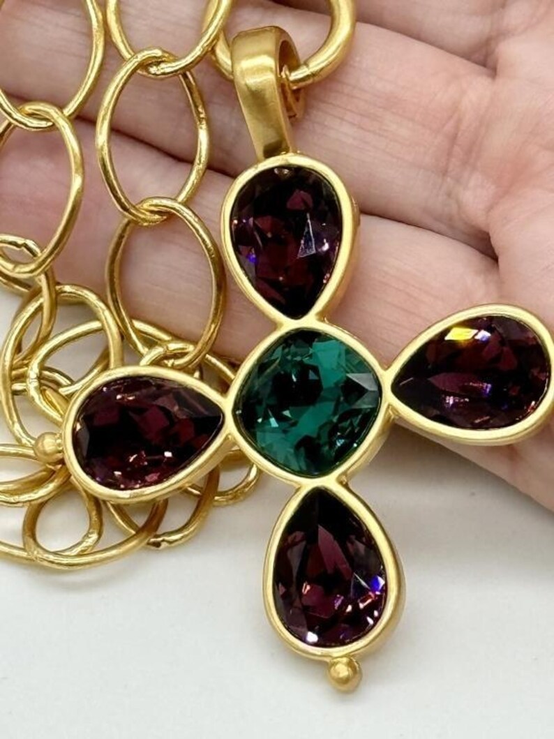 May include: A gold chain necklace with a pendant featuring a four-leaf clover design. The clover is made of four teardrop-shaped stones, two purple and two green.