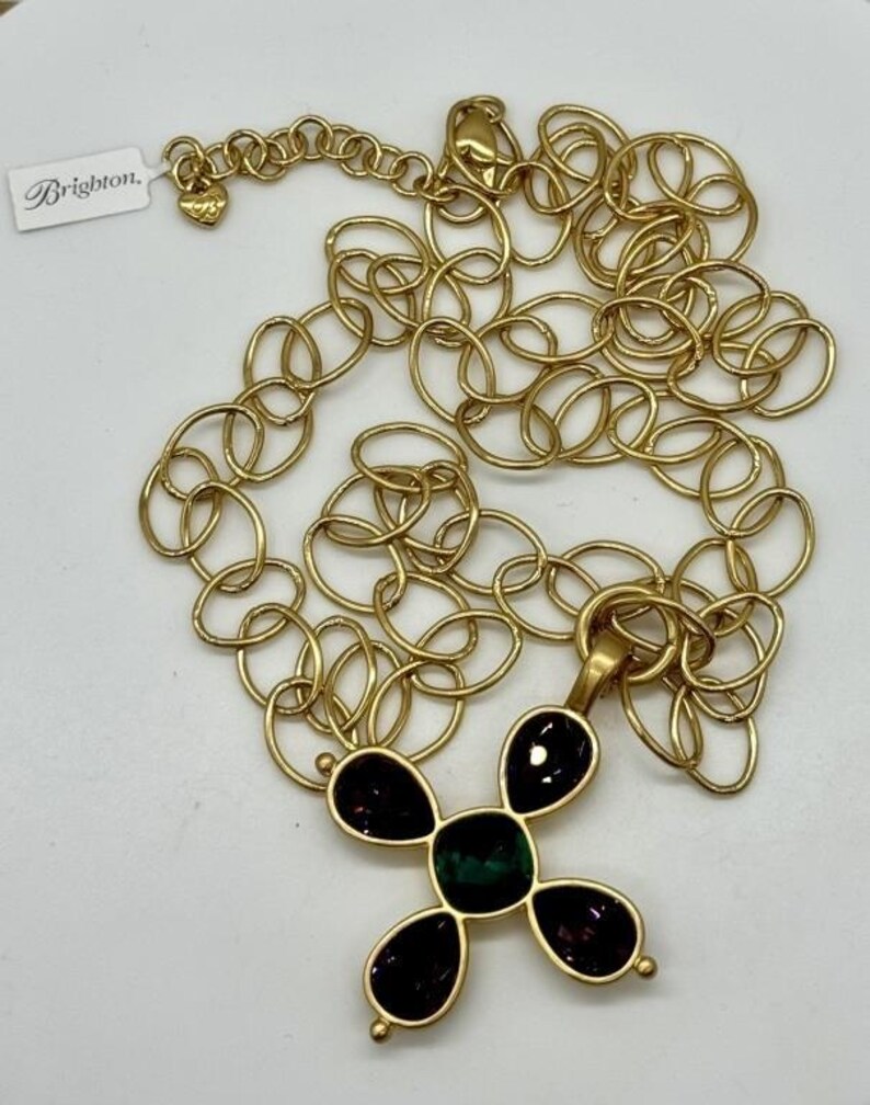 May include: Gold chain necklace with a pendant in the shape of a cross. The cross is adorned with five stones, four purple and one green.