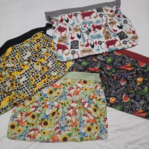 May include: Four fabric aprons with distinct patterns. One features farm animals and windmills on white, one has yellow sunflowers on black and white gingham, one displays colorful gnomes and sunflowers on light green, and one shows produce on black.