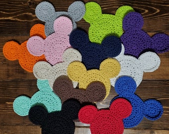May include: A collection of twelve crocheted coasters shaped like mouse heads in various solid colors including red, orange, yellow, green, blue, purple, pink, black, brown, white, gray, and light blue, arranged in a cluster on a dark wood surface.