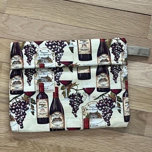 Può includere: Una pochette rettangolare a tema vino. Il tessuto beige è stampato con immagini di bottiglie di vino, grappoli d'uva e bicchieri nei toni del rosso, marrone e viola. Un piccolo passante in tessuto beige è attaccato al lato.