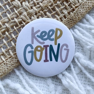Puede incluir: Un botón blanco con el texto "Keep Going" en letras de colores. Las letras son de varios colores, incluyendo rosa, azul, verde y amarillo. El botón está sobre una superficie blanca y peluda.