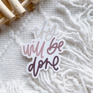 May include: A sticker with the text "thy will be done" in a pink and white color scheme. The sticker is on a white fuzzy surface.