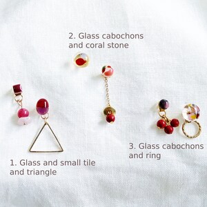 May include: Three pairs of earrings with different designs. The first pair features a pink and red glass tile, a small pink stone, and a gold triangle. The second pair has a red and white glass cabochon with a gold chain and a red stone at the end. The third pair has three red glass cabochons, a purple glass cabochon, and a gold ring.