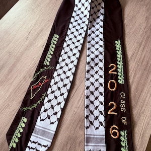 May include: A graduation stole with a black and white checkered design, with the gold text "CLASS OF 2026". The stole also has green and gold embroidery, and a map of Palestine with Arabic text.