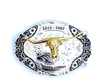 May include: Silver and gold-tone metal belt buckle with a longhorn steer design. The buckle has a decorative border with the text "2022-2082" at the top and "LUCKY COWBOY" at the bottom. A western-themed accessory.