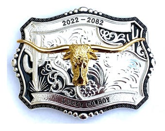 May include: Silver and gold-tone metal belt buckle with a longhorn skull design. The buckle features a gold-colored longhorn skull in the center, with the text "LUCKY COWBOY" and the years "2022-2082". The buckle has a black border and a decorative pattern.