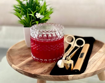 May include: A red candle in a clear glass jar with a diamond pattern. The candle is on a wooden tray with a black cloth and gold candle accessories.