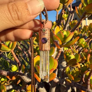 May include: A copper-colored pendant necklace featuring a clear crystal and three gemstones: green, purple, and clear. The pendant hangs from a brown cord, set against a backdrop of green and yellow foliage.